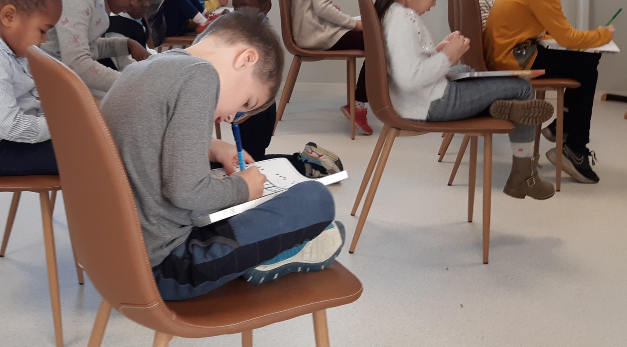 Barn sitter på spredte stoler, skriver på ark.