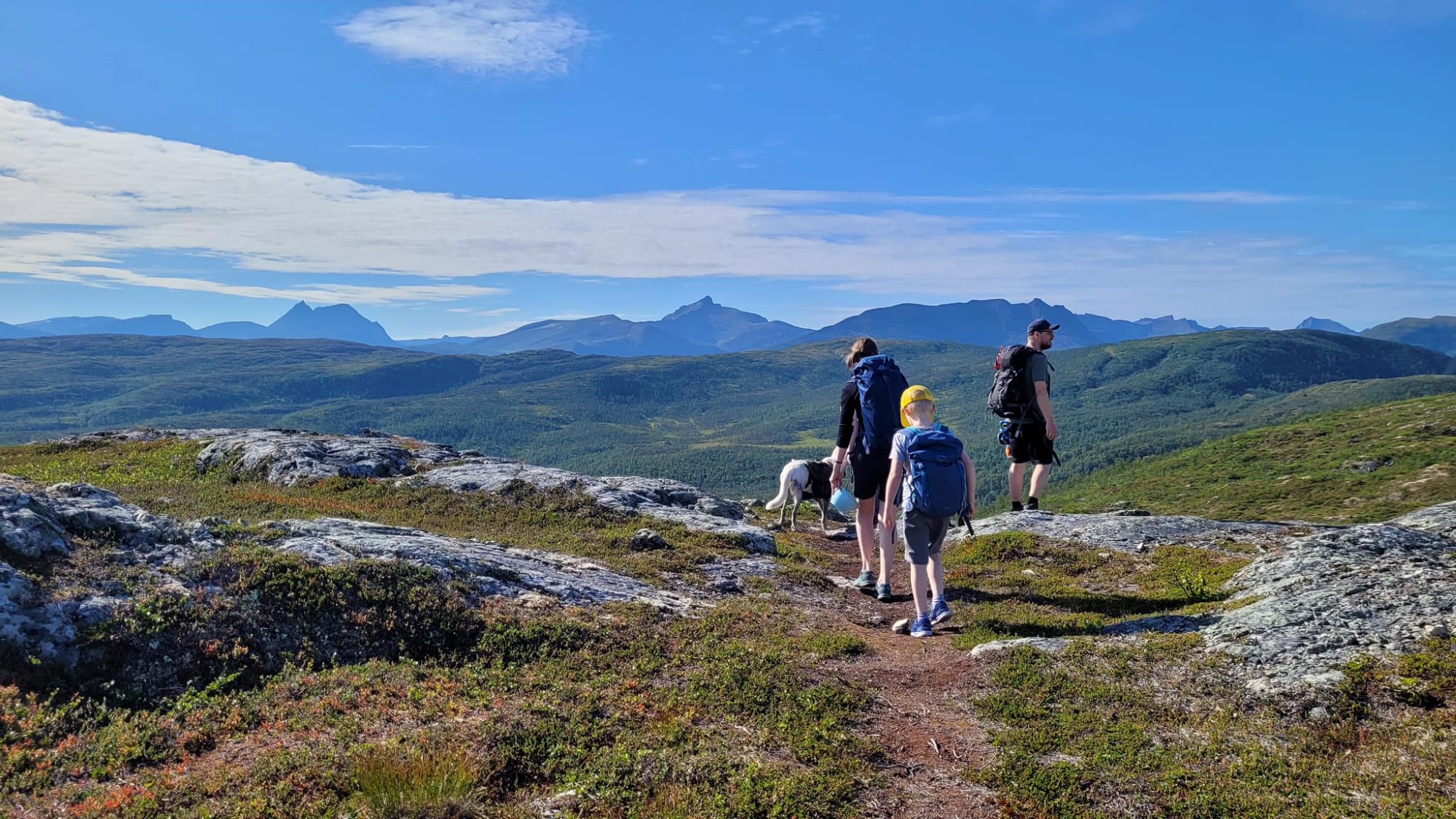 To voksne, et barn og en hund som går i fjellet med fin utsikt til fjelltopper