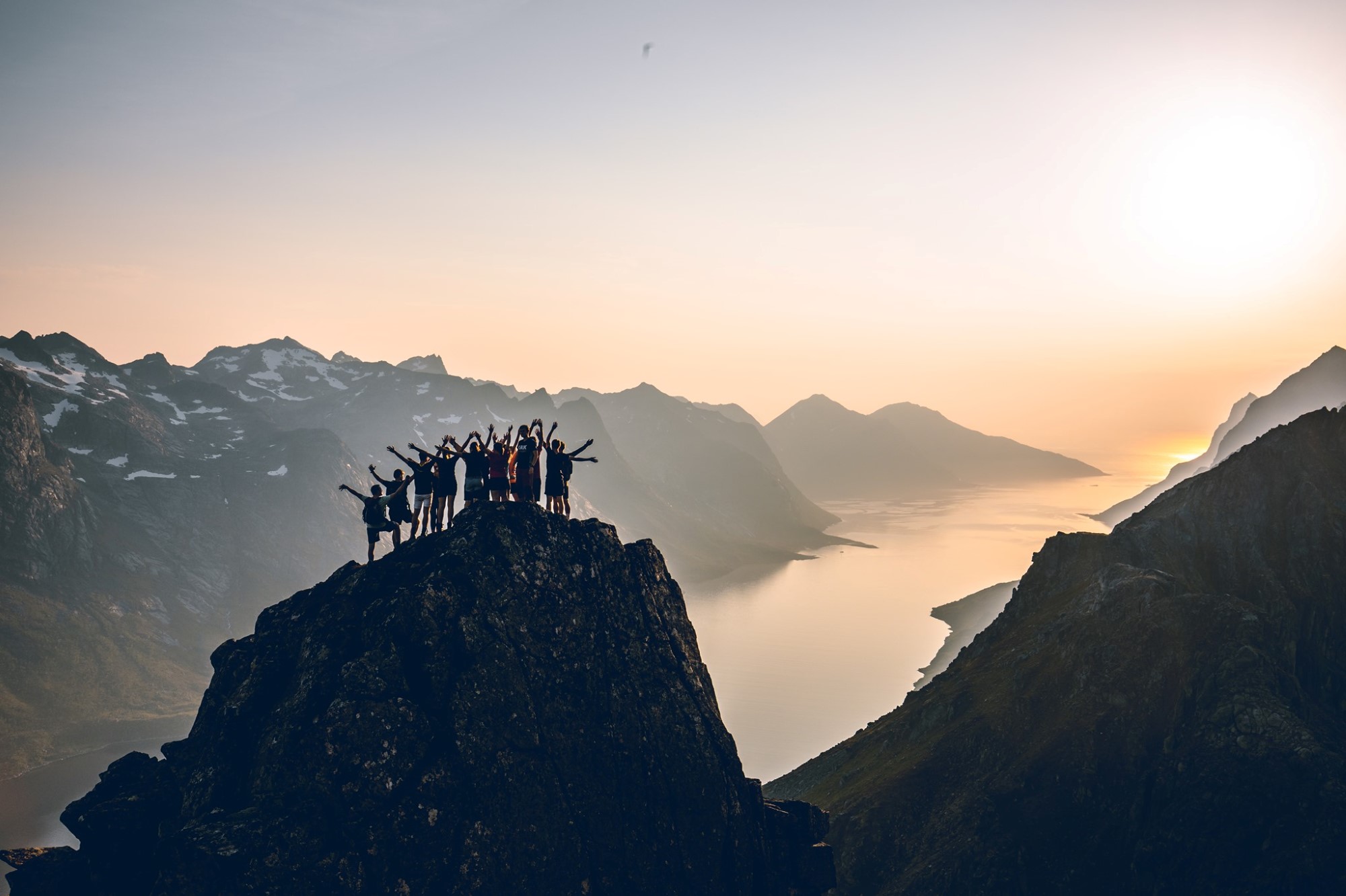 En gruppe mennesker står på en fjelltopp og ser mot sola utover en fjord med høye fjell