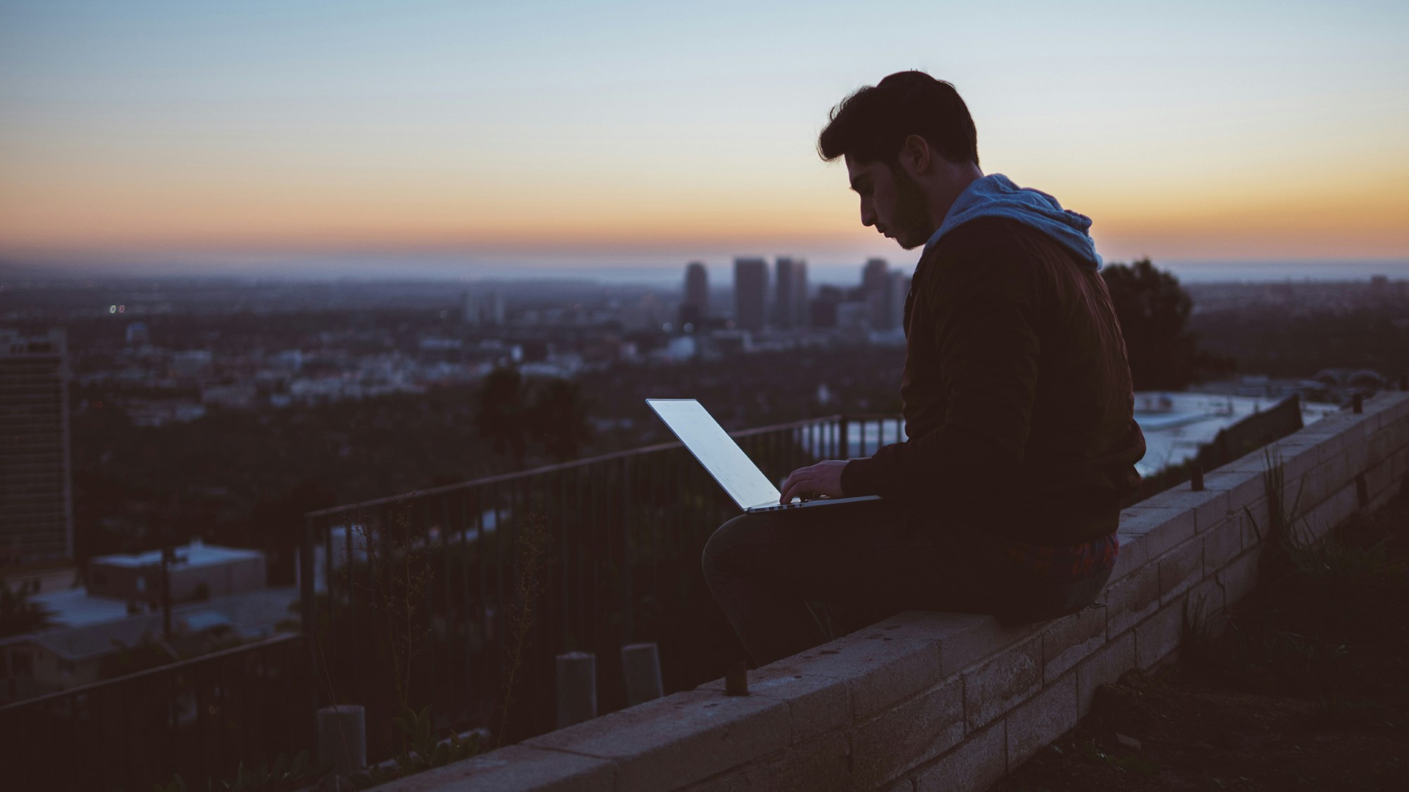 Mann sitter på tak i solnedgang og arbeider på PC